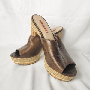 May include: Brown leather platform sandals with open toes. The sandals have a cork wedge heel and a woven jute platform. The brand name "PRADA" is visible on the insole.