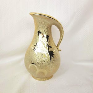May include: A gold-toned ceramic pitcher with a textured, speckled surface. The pitcher features a smooth, reflective gold rim and handle, with a stylised black and gold graphic design of a fish on the front and a crescent shape near the base.