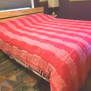 May include: A red and pink patterned blanket with a fringed edge is draped over a bed. The blanket features alternating stripes and square patterns in varying shades of red and pink, creating a textured appearance.