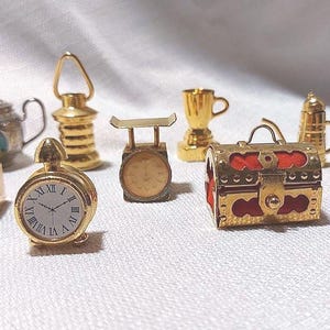 May include: A selection of small, gold-coloured ornaments. These include a clock, coffee grinder, teapot, lantern, scales, trophy, chest, coffee pot, and cannon. The clock face displays Roman numerals. The chest has red lining.