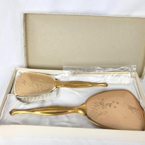 May include: A vintage vanity set in a box, featuring a brush and a hand mirror with gold-toned handles and floral designs. The set includes a comb. The brush and mirror have a light pink surface.
