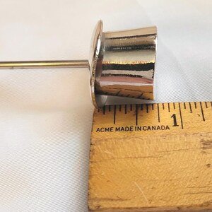 May include: A shiny, silver-coloured metal object with a cylindrical body and a flat, circular top. The object is next to a wooden ruler, with the measurement at approximately 2.5 cm.