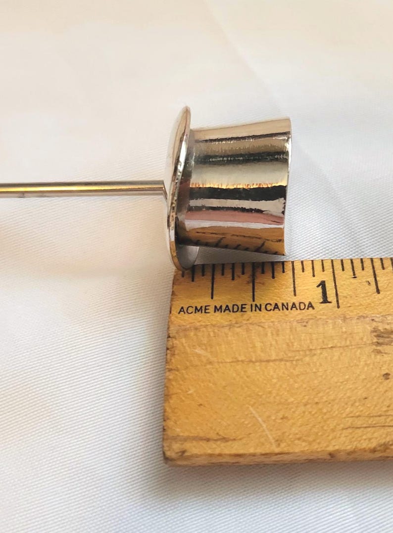 May include: A shiny, silver-coloured metal object with a cylindrical body and a flat, circular top. The object is next to a wooden ruler, with the measurement at approximately 2.5 cm.