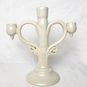 Peut inclure: Un candélabre en céramique de couleur crème doté d'un pilier central et de deux bras latéraux incurvés vers l'extérieur. L'objet possède trois bougeoirs, chacun avec une petite pointe à la base, et des ornements en forme de volutes en bas.