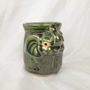 May include: A small, green ceramic mug with a textured surface and a sculpted, stylized face. The mug has a dark green glaze with lighter green accents and small, yellow-orange eyes. The mug is cylindrical with a slightly flared top.