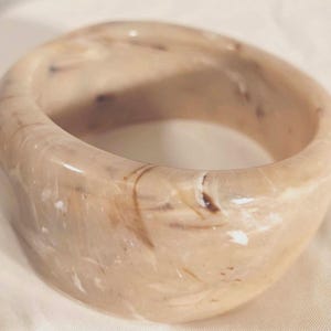 May include: A wide, circular bangle bracelet featuring a smooth, polished finish with a marbled pattern in shades of beige, cream, and dark brown. The thick, rounded band has a glossy surface that reflects light, creating a continuous, swirling effect.