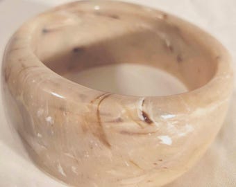May include: A wide, circular bangle bracelet featuring a smooth, polished finish with a marbled pattern in shades of beige, cream, and dark brown. The thick, rounded band has a glossy surface that reflects light, creating a continuous, swirling effect.