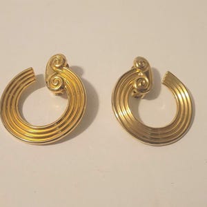 Puede incluir: Un par de pendientes de metal en tono dorado con un diseño de aro abierto circular y líneas paralelas estriadas. Cada pendiente está rematado con un motivo decorativo de doble espiral en el cierre, creando un aspecto estructurado.