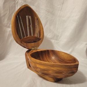 May include: A polished wooden nut-shaped bowl with a hinged lid that opens upward. The interior of the lid features a built-in rack holding a metal nutcracker and two metal picks. The exterior of the bowl has a textured, dimpled surface.