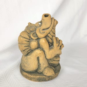 May include: A tan, textured ceramic sculpture of a seated creature with a prominent snout, large ears, and a rounded body. The figure is positioned on a circular base with vertical etched lines, featuring a hollow opening at the top of the head.