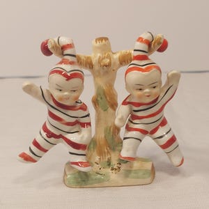 May include: Two porcelain figurines of children dressed in red and white striped outfits, standing on a green base with a brown tree trunk in the center.