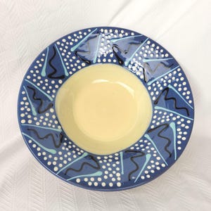 May include: A ceramic bowl with a wide, flat rim featuring a blue background decorated with white polka dots, light blue triangular shapes, and black wavy lines. The centre of the bowl has a smooth, solid pale yellow interior surface.