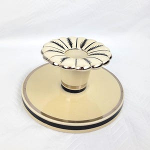 May include: A cream-coloured ceramic candle holder with a wide circular base and a scalloped top rim. The piece features thin black painted accents, including delicate feather-like patterns on the scalloped edge and a bold black band around the base.