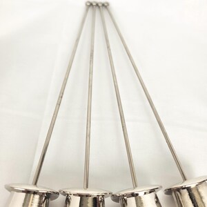 May include: Five silver cocktail picks are displayed. Each pick features a small, cylindrical base and a long, thin stem, culminating in a rounded tip. The picks are set against a white backdrop.