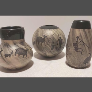 Peut inclure: Trois récipients en céramique avec une finition marbrée grise et noire. Chaque pièce présente des bords noirs et des silhouettes sombres de chèvres et de personnages. L'ensemble comprend un pot arrondi, un vase sphérique et un vase haut.