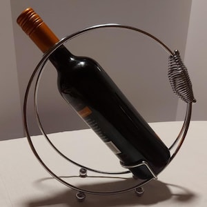 May include: A silver metal wine bottle holder with a circular design. The holder has a spring-like detail on one side and a bottle of red wine is resting inside.