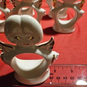 May include: Set of six white porcelain angel napkin rings with gold accents. Each ring features a detailed angel design with wings and a halo. The rings are approximately 2 inches in diameter.