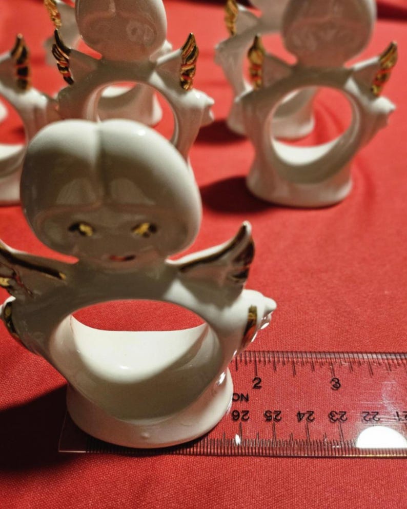 May include: Set of six white porcelain angel napkin rings with gold accents. Each ring features a detailed angel design with wings and a halo. The rings are approximately 2 inches in diameter.