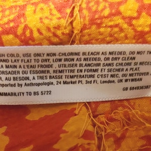 May include: A white care label with text in English and French, sewn onto a patterned orange and yellow fabric. The label provides washing instructions and flammability details. The fabric has an intricate design.
