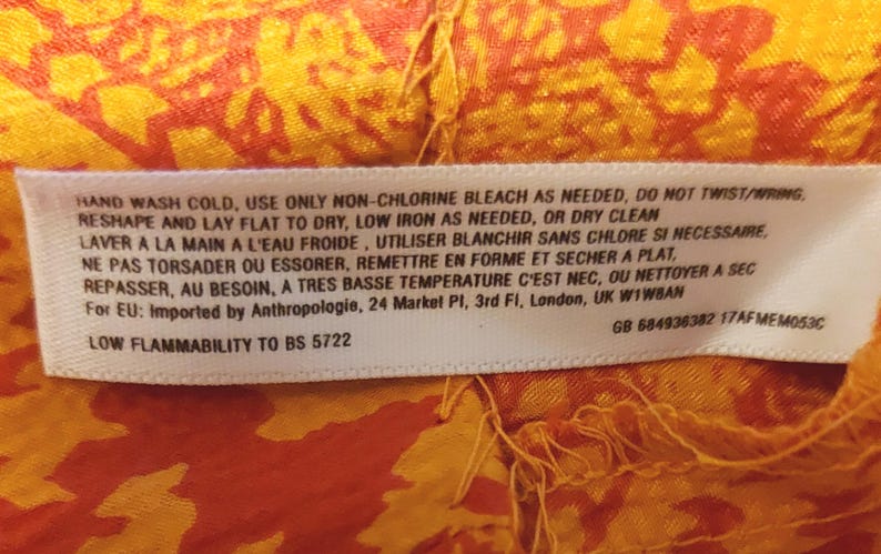 May include: A white care label with text in English and French, sewn onto a patterned orange and yellow fabric. The label provides washing instructions and flammability details. The fabric has an intricate design.