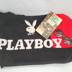 May include: A black and red towel featuring the white Playboy rabbit logo and the word PLAYBOY in large white letters. A small book titled Playboy's Party Drinks and a pair of dark-tinted sunglasses are placed on top of the towel.
