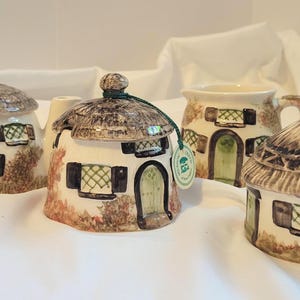 May include: A collection of ceramic cottage-shaped items, including a teapot, a sugar bowl, and a creamer. The pieces feature thatched roofs, small windows with green lattice, and brown accents. The items are painted in cream, brown, and green.