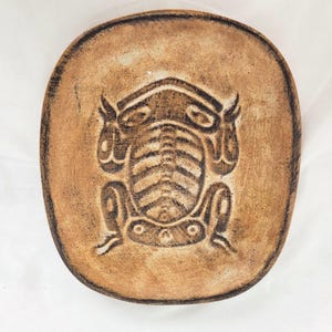 May include: A handmade, oval-shaped ceramic dish with a frog design. The dish is a light brown color with a darker brown border and frog illustration. The frog is depicted in a stylized, tribal art form. The dish is likely used for decorative purposes.