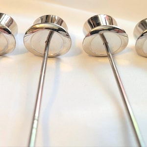 May include: Four silver cocktail picks with a rounded top and a long, thin stem. The picks are arranged in a row against a white background. The metal has a shiny, reflective surface. The picks are for serving appetizers.