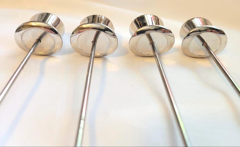 May include: Four silver cocktail picks with a rounded top and a long, thin stem. The picks are arranged in a row against a white background. The metal has a shiny, reflective surface. The picks are for serving appetizers.