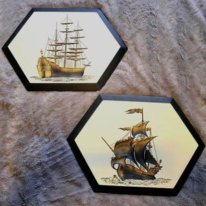 Peut inclure: Deux tableaux muraux hexagonaux représentant des illustrations de voiliers anciens. Les navires sont représentés en or, marron et noir sur un fond crème. Chaque pièce a un cadre noir, créant un thème nautique classique.