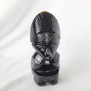May include: A polished black stone figurine carved into the shape of a stylised human figure. The sculpture features a prominent, fan-shaped headdress with vertical grooves, a cross-hatched headband, and simplified facial features on a smooth surface.