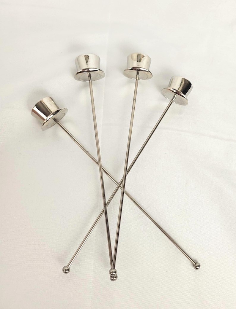 May include: Four silver cocktail stirrers. Each stirrer has a miniature top hat design at the top and a small ball at the bottom. The stirrers are set against a white background.