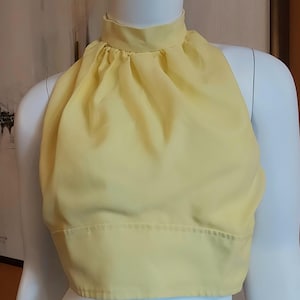 May include: A pale yellow sleeveless crop top featuring a high mock neck collar with gathered fabric detailing at the neckline. The garment has a wide, structured band at the waist and a smooth, solid-coloured finish, displayed on a white mannequin.