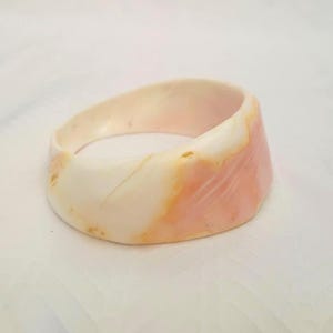 May include: A wide bangle bracelet crafted from smooth, polished material featuring a natural marbled pattern. The surface displays a blend of creamy off-white, soft peach, and pale pink tones with subtle, organic veining throughout the curved structure.