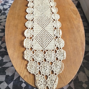May include: Cream-colored, hand-crocheted table runner. The runner features a central diamond pattern and a scalloped edge with floral motifs. A tassel hangs from one end. The runner is displayed on a wooden oval table.