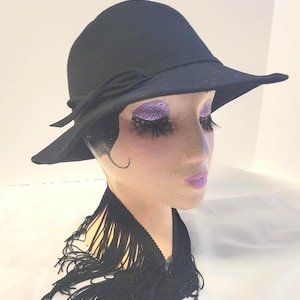 Peut inclure: Un chapeau en feutre noir avec un bord large légèrement incliné vers le bas et un nœud décoratif sur le côté. Le chapeau est présenté sur une tête de mannequin avec du fard à paupières violet pailleté, de longs cils et un collier à franges noir.