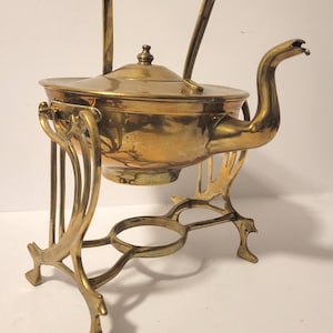 May include: A vintage brass tea kettle with a lid and a curved spout. The kettle sits on a decorative stand with an open ring and ornate legs. The handle is woven.