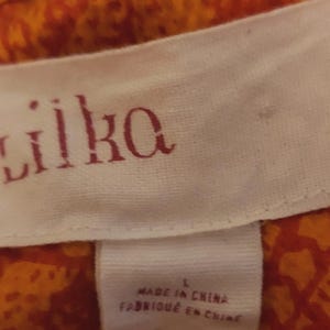 May include: A close-up of a white fabric label with the brand name "lilka" in red lettering. The label is attached to orange and red patterned fabric. The label also includes the text "MADE IN CHINA" and the size "L".