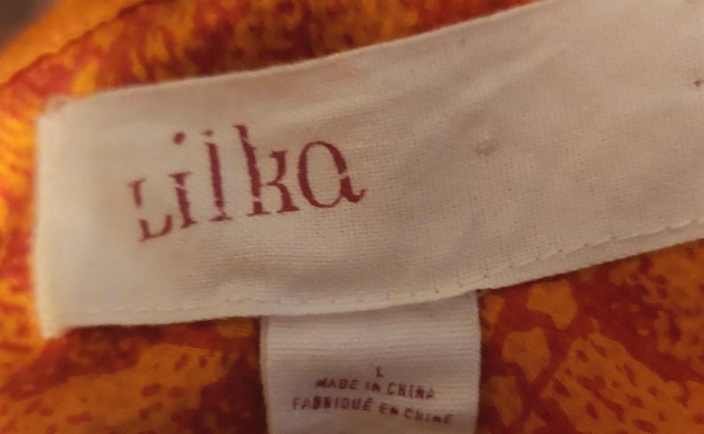 May include: A close-up of a white fabric label with the brand name "lilka" in red lettering. The label is attached to orange and red patterned fabric. The label also includes the text "MADE IN CHINA" and the size "L".