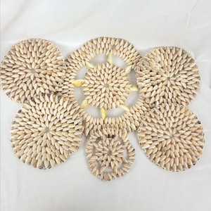 May include: A decorative set of coasters and a centerpiece, all crafted from small, cream-coloured seashells. The arrangement includes circular and petal-shaped elements, creating a natural, coastal aesthetic. The centerpiece has a diameter of approximately 15 cm.