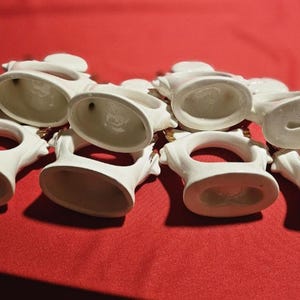 May include: Set of seven white ceramic napkin rings with gold accents. Each ring features a raised design and a circular opening for a napkin.