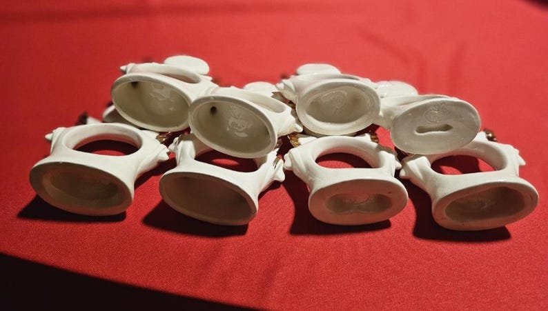 May include: Set of seven white ceramic napkin rings with gold accents. Each ring features a raised design and a circular opening for a napkin.
