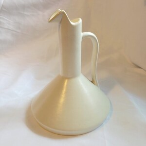 May include: Cream-colored ceramic pitcher with a tall, slender neck and a flared base. The pitcher has a curved handle and a spout with a slight gold accent. The design is simple and elegant, suitable for serving beverages or as a decorative item.