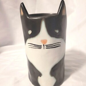 May include: A ceramic tumbler shaped like a cat with black and white markings. The vessel features two pointed ears at the top, a white face with black eyes, a small peach-coloured nose, and black painted whiskers on a white muzzle area.