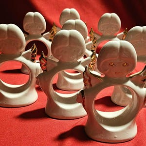May include: Set of ten white ceramic angel napkin rings with gold accents. Each ring features a sculpted angel with wings and a halo.