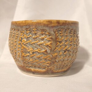 May include: A ceramic cup featuring a textured exterior with a repeating chevron pattern. The surface displays a blend of warm amber, brown, and muted grey glazes, with a smooth, rounded rim and a slightly tapered base design.