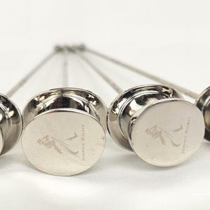 May include: Four silver cocktail picks with round, flat tops. Each top features an etched logo and the words "Johnnie Walker". The picks are arranged in a row, with the stems pointing upwards.