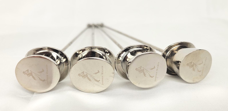 May include: Four silver cocktail picks with round, flat tops. Each top features an etched logo and the words "Johnnie Walker". The picks are arranged in a row, with the stems pointing upwards.