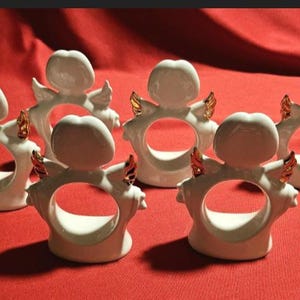 May include: Set of eight white ceramic napkin rings with gold accents. Each ring features a stylized angel design with wings and a halo.