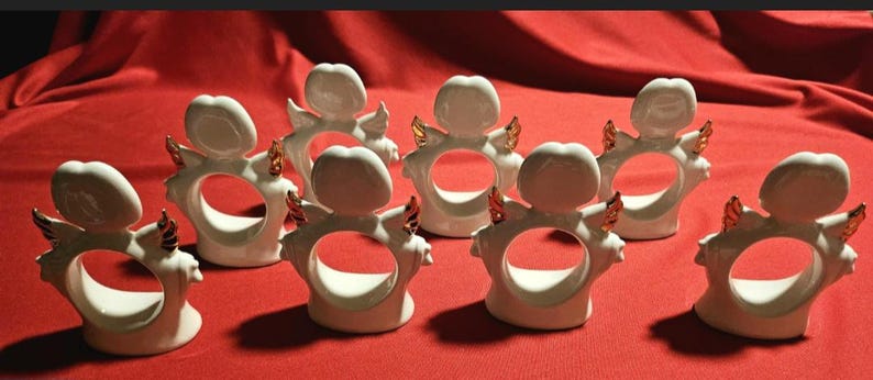 May include: Set of eight white ceramic napkin rings with gold accents. Each ring features a stylized angel design with wings and a halo.